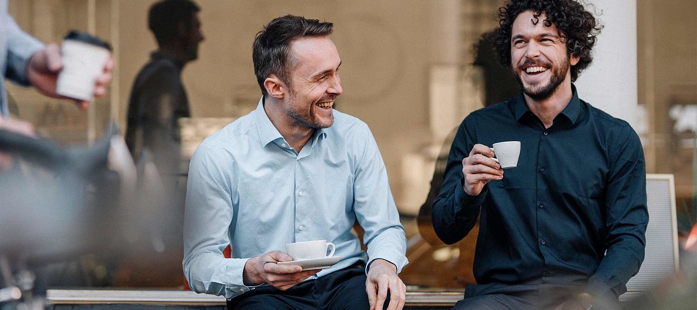 zwei Männer lachen und trinken Kaffee sitzend vor einem Fenster